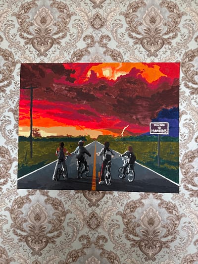 A painting depicts four figures on bicycles riding down a road toward a dramatic, fiery sunset. The sky is filled with vibrant reds, oranges, and yellows, with dark, ominous clouds and a streak of lightning. A sign on the right side of the road reads "WELCOME TO HAWKINS." The figures on bicycles are silhouetted against the bright sky, and their clothing appears to be dark. The road has a yellow dividing line and white edges. Green fields flank the road, and a utility pole stands on the left. The painting is displayed against a backdrop of light blue wallpaper with an ornate damask pattern in shades of gray and brown. This scene is not located in Kondopoga, Russia; it is a depiction of the fictional town of Hawkins from the television series "Stranger Things."