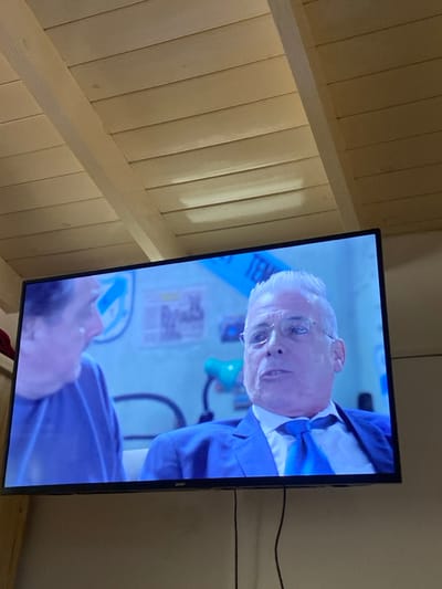 A television screen displays a scene with two men, one older with white hair and glasses wearing a blue suit with a bow tie, and another with dark hair and a blue shirt. A medical light and poster are visible in the background. The television is slightly askew against a white wall, with visible wires. To the left, a wall made of light-colored wood planks extends diagonally.
