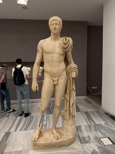 The image depicts a marble statue of a male figure, likely ancient Greek or Roman, displayed in a museum. The statue is life-sized and depicts the man nude, with a drapery draped over his left shoulder and arm, and held in his right hand. The statue is missing its lower legs, with the remaining stumps resting on a stone pedestal. The statue is set against a grey wall with modern lighting fixtures visible on the ceiling. In the background, two men are observing exhibits, one wearing a green cap and the other a white shirt and black backpack. The floor is made of marble tiles with a grey and white veined pattern. The statue is in a well-lit museum setting. The location is the Heraklion Archaeological Museum in Heraklion, Greece, a prominent museum housing artifacts from Minoan civilization and other periods of Cretan history.