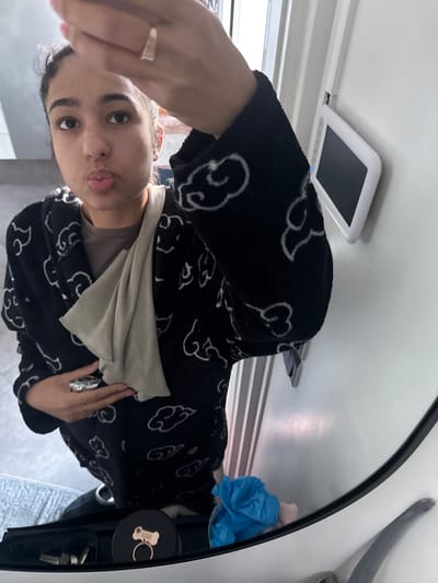 A single young female individual is depicted in an indoor setting, taking a mirror selfie. The scene is likely a private residence in Limeil-Brévannes, France. The female has dark hair, dark eyes, and a light brown complexion, and is looking directly at her reflection with a pursed-lip expression. She is wearing a black, plush robe or hooded garment patterned with white clouds, over a plain brown top. A light tan fabric is draped across her chest and left shoulder. Her right arm is raised, holding the device capturing the image, revealing a gold-colored ring on her right ring finger. Her left hand holds a small, metallic-wrapped object near her chest, with a silver-colored ring visible on her left ring finger. The background, reflected in the mirror, shows a white wall with a mounted rectangular white electronic device featuring a dark screen, possibly an intercom or control panel. Below the subject's reflection, on a visible surface, a black round container with a gold dog bone keyring, a crumpled light blue plastic bag, and other indistinct small objects are present. The floor appears tiled.