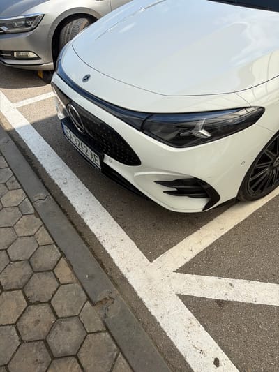 The image displays the front section of a white Mercedes-Benz sports car, likely a Mercedes-AMG GT, parked outdoors in Byala, Bulgaria, under bright daylight conditions. The car is angled, showcasing its sleek right headlight, distinctive black grille with a prominent Mercedes-Benz emblem, and a part of its multi-spoke black wheel. Reflections of what appears to be an overhead structure are visible on the car's glossy white hood. A minor scuff or scratch is present on the front bumper above the right air intake.

A European-style license plate, bearing the alphanumeric sequence "EN 3262 AE", is affixed below the grille. The front-left section of another silver or grey car is partially visible behind and to the left of the white Mercedes, suggesting a parking lot setting.

The ground beneath and around the cars is dark asphalt, marked with thick, weathered white lines that form a corner or chevron pattern, indicative of parking space delineation. A concrete curb separates the asphalt from an adjacent area paved with grey hexagonal blocks. The strong contrast and clear shadows suggest sunny weather.
