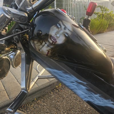 The image shows a close-up of a custom motorcycle's fuel tank, which features an airbrushed design of a woman's face with striking blue eyes and blood dripping from her mouth. The motorcycle is parked outdoors on a paved surface, possibly a sidewalk or driveway, with a white metal fence and green foliage visible in the background. The sun is casting warm light on the scene, reflecting off the motorcycle's glossy black paint. The motorcycle appears to be a cruiser style, with chrome accents and custom detailing. The surrounding environment suggests a sunny day in a residential or urban area. There are no people present in the immediate vicinity.