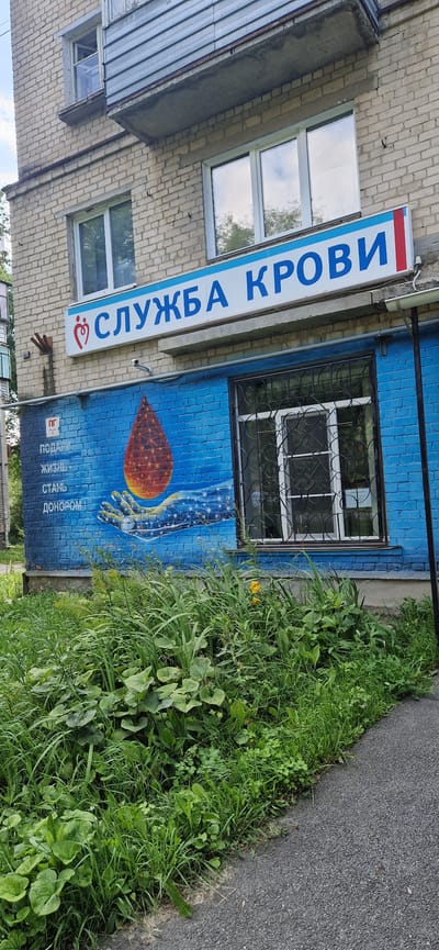 This image shows the exterior of a building in the city of Miass, Russia, which houses a blood donation service. The building has a brick facade and several windows. A prominent sign above a ground-floor window reads "СЛУЖБА КРОВИ" (Blood Service) in blue letters with a red and blue stripe on the right. Below the sign, a mural on the blue brick wall depicts a hand holding a glowing droplet of blood, symbolizing donation. Text on the mural reads "ПОДАРИ ЖИЗНЬ. СТАНЬ ДОНОРОМ!" (Donate Life. Become a Donor!). The surrounding area is overgrown with grass and weeds, with a paved pathway visible in the foreground. No people are visible in the scene.