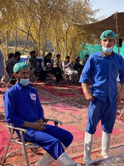 An outdoor scene in Sard-E 'olya, Afghanistan, shows two men in blue uniforms, light green hairnets, face masks pulled below their chins, and white rubber boots in the foreground, positioned on a red and black patterned carpet. The man on the left is seated in a silver metal folding chair, hands clasped, with a white badge displaying "364" and Farsi/Dari text pinned to his chest. The man on the right stands, facing forward with hands in his pockets. In the mid-ground and background, approximately ten other men are seated on similar folding chairs across the carpet, dressed in civilian attire, observing. The environment features numerous deciduous trees with yellow autumn foliage and some visible red fruits (likely pomegranates) under a clear sky. A patterned fabric canopy is partially visible in the upper right, and a light blue/green tarp is visible in the mid-background. The ground is entirely covered by multiple interconnected red and black patterned rugs.