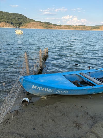 The image depicts a tranquil scene at a reservoir, likely the Yovkovtsi Reservoir, near Gniazdovo, Bulgaria. A vibrant blue rowboat, inscribed with Cyrillic lettering "МУСАЙБЕ 2020", rests partially on a sandy shore. The boat contains a coil of rope and a pair of oars, suggesting recent or planned activity. Adjacent to the boat, a fishing net is draped over a series of weathered wooden posts driven into the water. In the background, a small white boat is anchored on the calm, blue water, with rolling, tree-covered hills forming the distant horizon under a partly cloudy sky. The overall impression is one of peaceful, rural life.