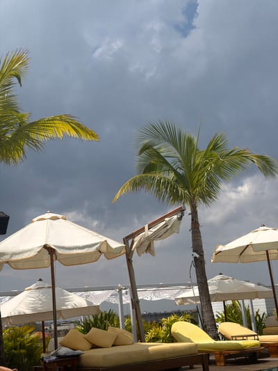 The image displays an elevated outdoor leisure area, likely a rooftop or poolside lounge, situated in Tibubeneng, Indonesia, under a heavily overcast sky with dark grey clouds. Several vibrant green palm trees are visible, with their fronds partially illuminated. The foreground features multiple open, cream-colored outdoor umbrellas, accented with gold trim and supported by wooden poles. Adjacent to these are several yellow-cushioned loungers and daybeds; one prominent daybed has three yellow throw pillows and a small wooden side table next to it. A partially folded umbrella canopy is attached to a wooden beam structure. The edge of a pool is discernible in the lower right. A white surveillance camera with a black cable is affixed to the trunk of a central palm tree. Additional green foliage and white architectural elements, possibly a railing or building, are visible in the mid-ground, with a distant cityscape or landscape on the horizon. No individuals are present in the scene.