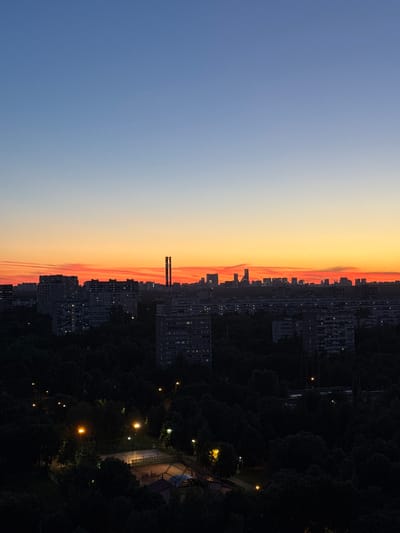 The image captures a twilight cityscape, likely in a residential area of Бордуки, Russia. The sky transitions from a deep blue overhead to a vibrant orange and pink sunset at the horizon. Silhouetted against this colorful sky are numerous apartment buildings, many with illuminated windows, suggesting inhabitants are active. In the foreground, a dense forest canopy is visible, punctuated by streetlights casting pools of yellow light on pathways and a recreational area that appears to be a sports court. The overall impression is one of a calm, urban evening setting.