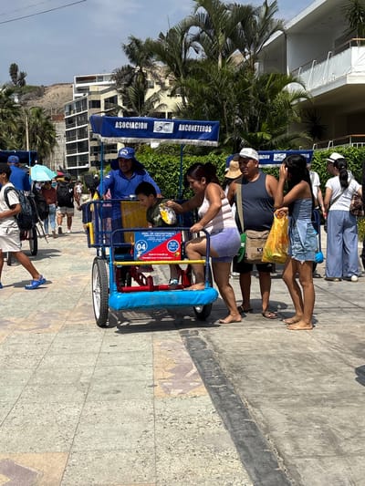 The image depicts a multi-person bicycle, occupied by a family of four, parked on a paved surface in Ancón, Peru. The bicycle is blue and yellow and has a sign that says "Asociación Anconeteros". The family consists of a mother, father, and two children. They are all barefoot. The mother is in the front, pedaling the bicycle. The father is behind her. The children are in the back. The pavement has a tiled design. In the background, buildings and palm trees are visible. Additional people are visible walking in the background. The sky is slightly overcast. The sun casts harsh shadows.