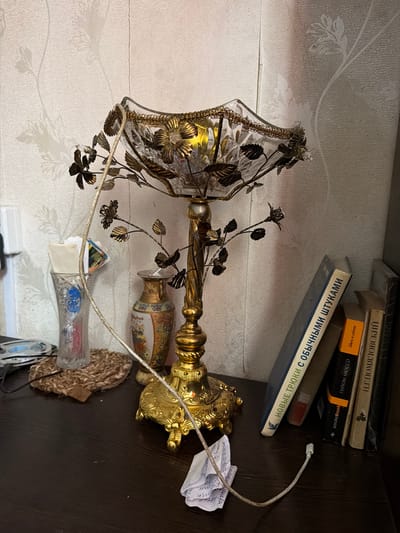 A still life image depicts a dimly lit interior scene, likely a bedroom or study, focusing on a decorative table lamp and surrounding objects. The lamp, made of ornate golden metal and clear glass with floral embellishments, sits on a dark wooden surface. A white power cord snakes from the lamp towards the foreground. To the left of the lamp, a clear glass vase holds a few small items, and behind it, a decorative vase with a patterned design. To the right of the lamp, a stack of books leans against the wall, their spines partially visible. The wall behind the objects features a textured wallpaper with a faint floral pattern, casting subtle shadows from the lamp. The overall impression is one of a quiet, perhaps personal, space. The image is not situated in Shepsi, Russia.