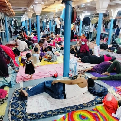 The image depicts a crowded indoor space, likely a passenger deck of a ferry or launch in Bhola, Bangladesh. Numerous people, including men, women, and children of various ages, are seen resting, sitting, and lying down on colorful mats and blankets spread across the floor. The setting is characterized by a series of blue-painted pillars with white decorative capitals, supporting a ceiling equipped with fluorescent lighting and fans.
In the foreground, a man lies on his side on a blue patterned mat, wearing blue jeans and a white sleeveless top. Nearby, other individuals are also resting on mats of various colors, including purple, pink, yellow, and red. A young child in a black dress walks barefoot across a pink mat in the mid-left. Many people are seen in relaxed postures; some are engaged with mobile phones, others converse in small groups, and several are sleeping. A woman in a red hijab and a light blue face mask is visible sitting towards the right.
Bags, backpacks, and personal belongings are scattered among the people. Visible objects include water bottles, one with the brand name "MOJO" clearly visible, and snack packets. In the background, clothes are seen hanging, suggesting a longer journey or temporary living arrangement. A large, dark, drum-like object is suspended from one of the blue pillars. The overall atmosphere suggests a period of travel and rest within a bustling but calm environment.
Visible text on a white mat in the foreground includes the Bengali words "এসো" (Esho, meaning "come") and "বৈশাখ" (Boishakh, a month in the Bengali calendar), repeated several times. The brand name "MOJO" is also visible on a water bottle. Other OCR-detected text like "পি ৬০৪" (P 604), "BIRIG", and "4010" is not distinctly identifiable in specific objects within the image but could be present on labels or distant items.