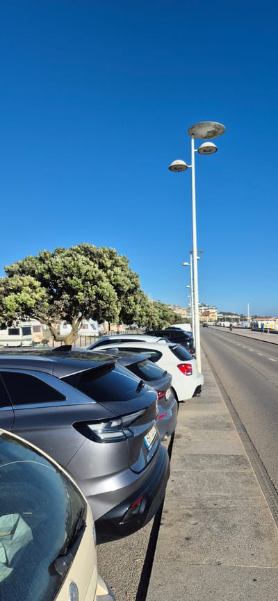 This image depicts a view along a street in Vila do Conde, Portugal, likely a coastal or promenade area, given the presence of palm trees and buildings suggestive of a resort town. The scene is dominated by a bright blue sky. In the foreground, a row of cars is parked along a sidewalk. The closest car is light beige, with its windshield and hood visible, showing some dust. Behind it is a dark gray SUV, notable for its modern design and taillights. Further down the line, a white SUV and other vehicles are parked.

To the left of the cars, a large, lush tree with dense foliage is prominent, casting shadows on the ground. Beyond the parked cars and the tree, a strip of sand or a beach appears to be present, followed by a line of buildings with balconies, suggesting residential or commercial establishments. Several tall lampposts, characteristic of a promenade or public walkway, are spaced along the right side of the image, lining the road.

The road itself is visible to the right, with traffic. People are seen walking and cycling on the sidewalk or promenade area adjacent to the road, enjoying the sunny weather. The overall impression is of a vibrant, public space in a Portuguese town, likely during daylight hours on a clear day.