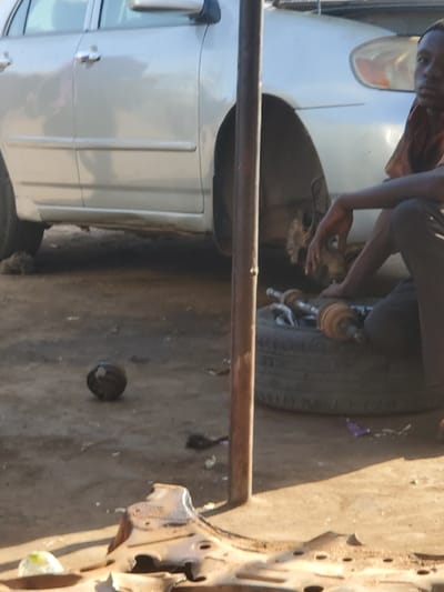 In Kaduna South, Nigeria, a daylight scene depicts an outdoor vehicle repair area. A silver sedan, identified as a Toyota Corolla, occupies the left side of the frame, with its front passenger-side wheel removed, exposing the brake caliper and hub assembly. A young male, wearing a rust-colored shirt and dark trousers, is seated on the unpaved, dusty ground in the mid-right. His right hand rests on his knee. Adjacent to him, a detached black car tire lies flat, supporting several metallic automotive components, including a drive shaft. A dark, vertical metal pole stands centrally. The immediate foreground shows a large, rusted, irregularly shaped metal car part and a dark spherical object on the ground. Scattered debris is present across the dirt surface. Shadows indicate direct sunlight.
