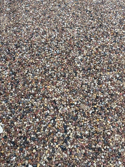 The image shows a close-up, overhead view of a surface covered in small, multi-colored gravel. The gravel consists of pebbles in shades of brown, grey, white, pink, and black. The texture is rough and uneven due to the varied shapes and sizes of the stones. There are no people, animals, or discernible objects other than the gravel itself. The lighting appears to be natural and overcast, casting soft shadows. The location is described as Капыльскі сельскі Савет, Belarus.