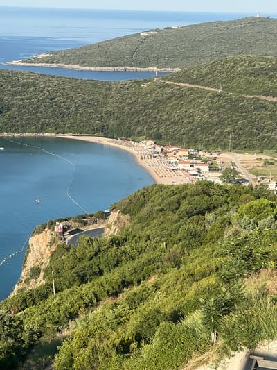 The image shows a panoramic view of a coastal area in Seoce, Montenegro. In the foreground, a steep, green, and heavily wooded hillside descends towards a body of water. A winding road is visible carved into the cliff face, with a few poles and some signage along its edge. The sea is a vibrant blue, with buoys marking off sections of the water.
Further out, a crescent-shaped beach is lined with numerous beach chairs and umbrellas, suggesting a popular tourist destination. Behind the beach, a cluster of buildings with red-tiled roofs forms a small village or resort. A road runs alongside the village, with several parked cars visible.
In the background, the landscape is dominated by rolling, tree-covered hills that extend towards the horizon. The sea stretches out to meet a clear blue sky. The overall scene depicts a sunny day at a scenic coastal location.