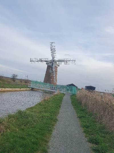 The image depicts a static rural scene featuring St Benet's Abbey Windpump near Ludham, North Norfolk, United Kingdom, under an overcast sky. A gravel path bisects the foreground, bordered by short green grass on the left and taller, dry reeds on the right. To the left of the path, a calm body of water, consistent with a drainage canal, flows. A light-colored metal bridge, partially obscured by green netting, spans this waterway, leading towards the base of a prominent brick tower windmill in the mid-ground. The windmill possesses distinctive skeletal metal sails. Behind the windmill, to the right, are several dark, low-lying structures. The sky is uniformly grey with diffused cloud cover. No individuals or active subjects are present.
