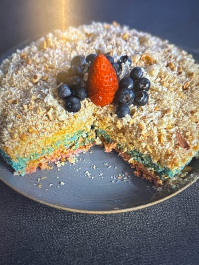A slice has been removed from a round cake which has layers of different colors, including blue, green, yellow, and pink. The top of the cake is covered in a crumbly topping, possibly nuts or streusel, and garnished with blueberries and a strawberry. The cake is presented on a dark gray plate with a light-colored rim. The plate sits on a dark, textured surface. The background is blurred and appears to be indoors, with a bright light source to the upper left. There is no discernible information in the image to suggest a location in Murcia, Spain.
