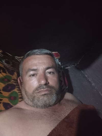 A medium-shot, low-light selfie of a middle-aged man with graying hair and a beard. He appears to be lying down, partially covered by a brown blanket. He is shirtless. He is against a wall, with colorful patterned fabric visible behind his head and to his left. There are no other people visible. The location is unknown.