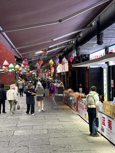 The image depicts an outdoor pedestrian street market located in Pengshui Miao and Tujia Autonomous County, China. The street surface is paved with irregular grey stone blocks. Overhead, a series of dark red retractable awnings are extended, supported by metallic frames. Beneath these awnings, numerous decorative lanterns, primarily teardrop or gourd-shaped and displaying various colors including pink, purple, yellow, green, and orange, are strung across the thoroughfare.
On the right side of the street, multiple market stalls are set up. The foreground stall features a white counter with red and black Chinese characters, including "鲜切水果捞" (Freshly Cut Fruit Salad) and "现榨果汁" (Freshly Squeezed Juice). Boxes containing goods are visible on and beside this counter. Further down the street, additional stalls display various food items, some under clear dome covers, against dark-colored shop fronts with illuminated signage featuring Chinese characters. One stall has a colorful graphic banner. A female individual, seen from behind, wearing a light-colored puffer vest, dark jeans, and a white apron, stands near the foreground stall, facing it. Behind her, a female vendor in a black top and apron stands behind another stall.
On the left side of the street, building facades are visible, with a prominent red wall partially visible above the awnings further down. This side also hosts various shops or stalls.
Approximately fifteen to twenty individuals are present on the street. In the mid-ground, a person wearing a dark long coat and a light-colored baseball cap is observed standing, holding and eating from a plate with a utensil. Adjacent to them, another person in a leopard-print jacket and brown skirt stands, facing slightly away. To their left, a person in a light-colored textured jacket and tan pants walks away from the camera. Further left, another individual in a black hooded jacket walks away. On the right side of the mid-ground, a person in a purple hoodie, grey pants, and blue sneakers stands, looking towards the camera. Pedestrians are observed walking in both directions along the street, and some are congregated near the stalls. The lighting appears to be natural daylight, with areas beneath the awnings providing shade, complemented by artificial light from stall signage.
