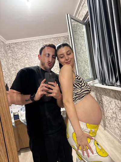 A man and a pregnant woman are taking a selfie in a room. The man is wearing a black t-shirt and black pants, and is holding a smartphone in his right hand. He is making a kissing face. The woman is wearing a zebra-print top and yellow pants with a cartoon character print. She is leaning towards the man and smiling. The room appears to be a bedroom, with a bed visible in the background. A window with dark curtains is also visible. The lighting is artificial. The image does not provide any specific information about the location in Tbilisi, Georgia.