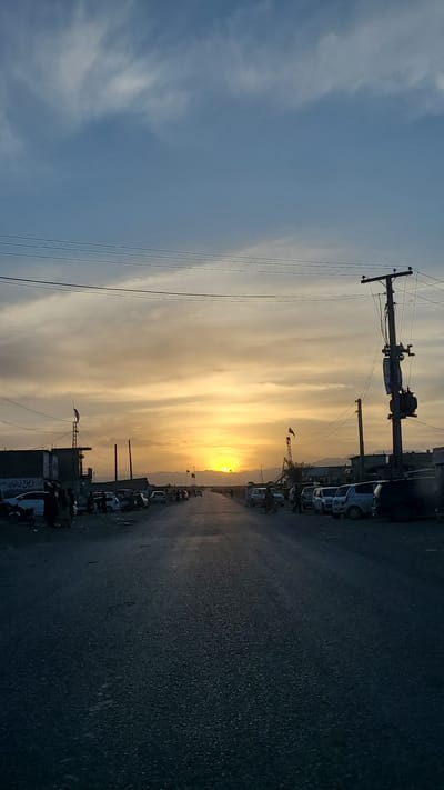 The image depicts a sunset scene along a street in Qilla Saifullah, Pakistan. Overhead power lines and poles are visible against the sky. Cars are parked along the right side of the street, and the road surface appears dark. The sun is setting over a distant horizon.