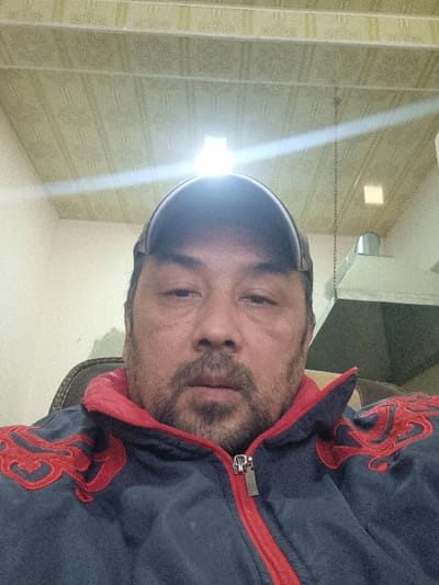 A man with dark hair and a stubbly beard is facing the camera, wearing a dark blue jacket with red trim and a dark baseball cap. The setting appears to be indoors, with a textured wall visible behind him. A bright light shines above, creating a glare. The photo is taken at an angle.