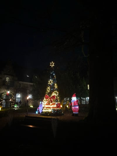 A nighttime scene depicts a festive outdoor Christmas display. A decorated tree, lit with stars and other holiday lights, dominates the center. An inflatable Santa Claus stands to the right of the tree. Behind the tree, a building with illuminated windows is visible. Further down the image, there are buildings with more Christmas decorations on them. The image is dark and captured from a low angle.