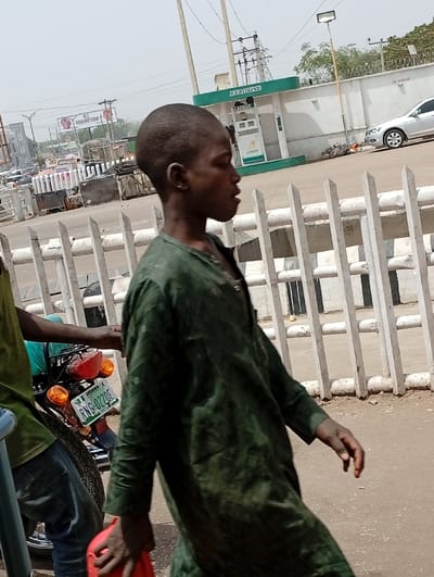 A young male with dark skin and a buzz-cut hairstyle walks from right to left in profile, his mouth slightly open, holding a red plastic jerry can in his right hand. He wears a dark green, long-sleeved garment. He is positioned in the foreground of an outdoor urban environment identified as Kofar Mazugal, Nigeria. To his left, partially visible, is another individual wearing a green top and blue jeans, standing next to a motorcycle. The motorcycle's rear, featuring red taillights, orange turn signals, and a Nigerian license plate "RNG-022Q5" from Kaduna State, is visible. A white metal barrier, consisting of vertical slats, separates the immediate foreground from the midground. Beyond this barrier, an "AZMAN KEROSINE" fuel station is visible with a green canopy and a white fuel dispenser. A silver sedan is parked near the fuel station. Overhead power lines and utility poles are present. Distant buildings, hazy sky, and a "NO SMOKING" sign on a wall are also visible in the background. The ground is a light brown, earthy surface.