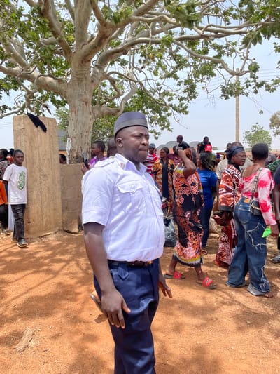 This outdoor photograph from Ladura Foron, Nigeria, captures a gathering of people under bright daylight. In the foreground, a man in what appears to be a uniform stands prominently, facing the viewer's left. He wears a white short-sleeved shirt with a crest on the left chest pocket, dark blue trousers, and a dark cap. His posture suggests observation or participation in an event.
Behind him, a group of individuals, predominantly women, are assembled. They are dressed in a variety of colorful and patterned traditional attire, with some also wearing jeans. One woman in the mid-ground stands out in a black dress adorned with orange floral patterns, wearing red sandals with visible text that appears to read "PUSH."
To the left of the frame, a large tree with light-colored bark and green foliage provides a natural canopy, its branches spreading across the upper left sky. The ground beneath is a reddish-brown, dusty earth, characteristic of the region. A structure made of unpainted mud or earth blocks is visible in the mid-ground, to the left of the assembled group. A young boy, wearing a white t-shirt with a distinct green design, stands next to this structure.
The clear sky and strong shadows indicate the time of day is likely mid-morning to early afternoon. The scene depicts a calm assembly, with individuals standing and looking forward, suggesting a community event or meeting. No explicit activity beyond gathering is immediately apparent, and the expressions on visible faces are largely neutral or attentive.