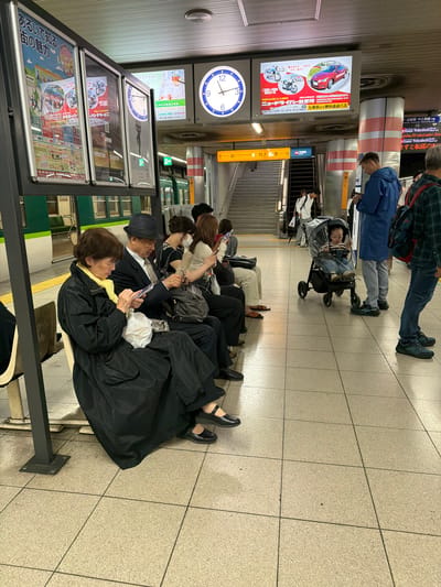 This image depicts an indoor subway station platform in Kyoto, Japan, identified by visible Japanese text and specific Kyoto references on posters.

On the left, a green and white subway train is visible through the platform windows. In the foreground, a row of individuals is seated on a beige bench. An elderly East Asian woman, wearing a black coat and a yellow scarf, is seated, engrossed in her white smartphone. A white plastic bag rests on her lap. To her right, an elderly East Asian man, dressed in a dark suit, light tie, and a dark fedora hat, is also seated and looking at his smartphone. A dark messenger bag is placed on the floor beside him. Further to the right, two additional East Asian women are seated, both occupied with their smartphones; one wears a white face mask.

In the middle of the tiled floor, a baby of indeterminate gender is positioned in a black stroller with a clear rain shield, looking towards the right. To the right of the stroller, a male, likely East Asian, wearing a blue hooded jacket and light grey pants, stands while viewing his smartphone. His feet are clad in dark athletic shoes. A male, likely East Asian, wearing a green and blue plaid shirt and carrying a dark backpack, is partially visible on the far right.

The station ceiling is fitted with white panels and fluorescent lighting. Above the platform, a large analog clock with Roman numerals, indicating approximately 12:15 or 12:20, is mounted on the wall. To the right of the clock, two digital display screens are active; the left screen advertises a driving school with images of a red car and motorcycles, while the right screen displays an interior scene with Japanese text and a phone number. A green illuminated exit sign is visible below the digital screens.

In the background, an escalator and a set of stairs lead upwards. A young East Asian male, wearing a white long-sleeved shirt and a black backpack, is seen ascending the stairs. Directional signage, featuring yellow and orange arrows, indicates "Exit" (出口) and "Subway" (地下鉄). The floor is covered with light beige-grey square tiles with dark grout lines. A cylindrical pillar with red and white stripes stands near the escalator.