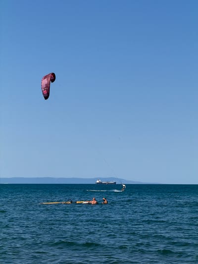 A wide shot captures a bright, sunny day at the coast of Atiya, Bulgaria, with a clear blue sky and calm, deep blue water. In the distance, a faint, hazy mountain range forms the horizon line. On the water, a lone kitesurfer glides across the surface, their kite a vibrant splash of red against the sky. Further out, a large cargo ship and a smaller vessel navigate the sea. In the foreground, two people are partially submerged in the water, one appearing to be resting on a yellow inflatable mat, while another person, wearing a green cap, looks towards the shore. The overall scene conveys a sense of leisure and outdoor activity on a tranquil sea.