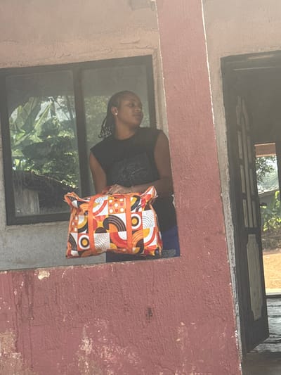 The scene depicts a female individual standing within an opening of a weathered, red-painted building in Aguata, Nigeria. The individual has dark skin, dark hair styled in braids or dreadlocks, and wears a dark, sleeveless top. She is positioned to face right, away from the camera, with her hands resting on a large, soft-sided bag. The bag features a vibrant geometric pattern in white, orange, red, brown, and black. Behind her, through a dark-framed window, lush green foliage is visible. To the right of the individual, a dark, partially open doorway reveals a sandy or dirt ground beyond. Strong sunlight illuminates the scene from the upper right, casting bright highlights on the red wall and the subject's hair.