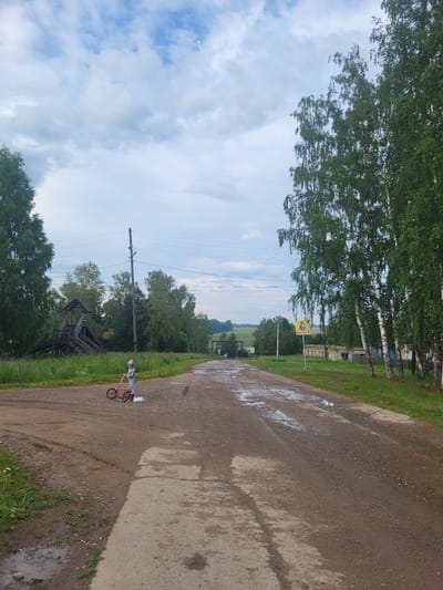 This media file depicts a rural scene in Zipunovo, Russia. A child, wearing a white and black patterned outfit and a helmet, is riding a small red bicycle on a wet, gravel road. The road is flanked by a grassy verge and tall trees with green foliage. In the background, there are more trees and a few buildings, suggesting a village or rural settlement. Power lines are visible stretching across the sky. There is a yellow road sign with a black symbol, possibly indicating a hazard or caution, on a pole to the left of the road. The sky is partly cloudy with patches of blue visible, and the overall lighting suggests it is daytime, likely in the afternoon. The presence of puddles on the road indicates recent rain. The scene evokes a sense of a peaceful, everyday moment in the countryside.