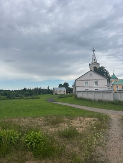 The image captures a serene, slightly overcast day in the village of Рассвет, Russia. In the foreground, a dirt path winds through a grassy area with tall weeds and some leafy green plants. The path leads towards a cluster of buildings, dominated by a light pink, two-story structure with a distinctive white brick wall in front of it. To the right of this building, a white and yellow bell tower, topped with a spire and a small cross, rises against the cloudy sky. Adjacent to it, another building with a green roof is partially visible. Further in the background, a line of trees creates a dark green backdrop, and to the left, a low bridge spans a body of water surrounded by vegetation. The overall scene suggests a quiet, rural atmosphere with traditional architecture.