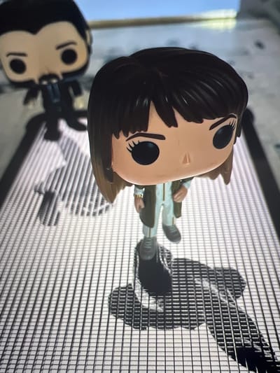 This is a close-up, high-angle, dimly lit photograph of two Funko Pop collectible figures. In the foreground, a female figure with dark brown bobbed hair, large black eyes, and light-toned skin is depicted wearing a white jumpsuit and a brown jacket. Her stance suggests she is walking. Behind her, and slightly out of focus, is a male figure with dark hair and a beard, also in a dark outfit. Both figures cast distinct shadows on a grid-like surface, suggesting they are on a grate or a textured floor. The background is blurred, showing a light-colored wall and a hint of blue sky. The image does not contain any information about the city of Mollet del Vallès, Spain.