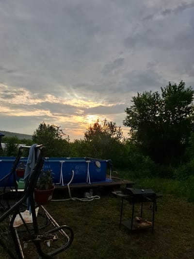 The image depicts a backyard scene in Sosnovka, Russia, at dusk. A large, blue Intex above-ground swimming pool is prominently featured, resting on a wooden platform. In the foreground, a dark metal swing set is visible, with a towel draped over its frame. A charcoal grill, also dark metal, stands to the right of the swing set, with firewood stacked on its lower shelf. The background is filled with lush green trees and vegetation, with a glimpse of a gentle hillside on the far left. The sky is overcast with dramatic clouds, through which rays of sunlight are breaking, creating a dramatic sunset effect. The overall atmosphere is peaceful and suggests an outdoor leisure setting.
