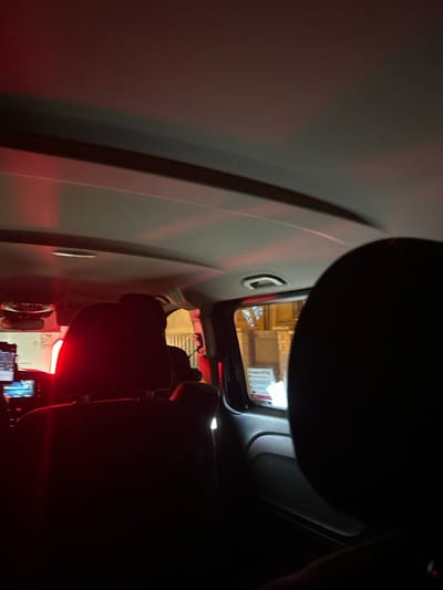 This interior shot, taken from a vehicle, shows the back of front seats and a glimpse out of the vehicle window. The interior lighting is dim, with red accents. The overhead sun visor is partially visible. Outside the window, a building facade is visible. In the foreground, a mobile phone sits on the armrest. The image seems to have been taken from the back seat of the vehicle.
