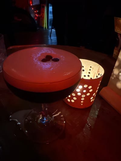 An eye-level, off-center shot captures an illuminated scene. A round, peach and black object with three small holes dominates the foreground, situated to the left of an empty wine glass. Behind it, an orange lantern with a dotted pattern casts light. The surfaces are dark, glossy, and reflect the light. The background is obscured, revealing only a hint of other elements.