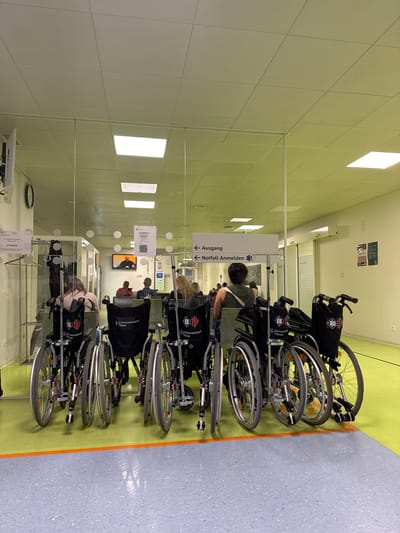The scene is an indoor corridor at the University Hospital Basel in Basel, Switzerland. A line of black wheelchairs with the university logo are parked along a green and blue patterned floor. Beyond a partial glass barrier, several people are seated. Overhead signage is visible, pointing to "Ausgang" (Exit) and "Notfall Anmelden" (Emergency Registration). A television screen displaying a person is visible on the far wall. The corridor is illuminated by rectangular ceiling lights. A clock and posters can also be seen on the wall.
