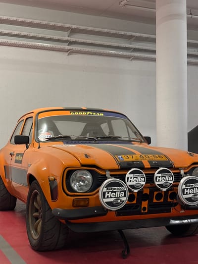 An orange Ford Escort Mk1 rally car is parked stationary, angled slightly towards the left, in an indoor facility in Bnei Brak, Israel. The vehicle features two black racing stripes running down its hood and roof, with yellow "GOODYEAR" lettering on a black strip across the top of the windshield and on the front left fender. An Israeli license plate "37-40-56" with Hebrew text "רכב אספנות" (Collector Vehicle) is mounted on the front. Four white circular HELLA auxiliary lights (two "Comet 500" and two "Rallye 3000") are prominently displayed on the front bumper. A "GULF" sticker is visible on the windshield. The car is equipped with wide tires on multi-spoke wheels, visible hood pins, and a black rectangular side mirror on the driver's side. Through the windshield, Sparco racing seats and a roll cage are discernible. The immediate environment consists of a smooth red floor, light grey concrete walls with some texture, and a portion of a white round concrete pillar visible on the right side of the frame. The ceiling above shows exposed utility conduits. No individuals are present in the scene.