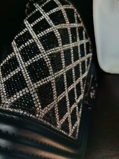 A close-up, slightly out-of-focus shot shows a black shoe with a diamond pattern. The diamonds are arranged in a grid and appear to be made of rhinestones. The shoe is on a dark surface, and a white object is visible to the right. The image does not provide any information about the city of Aksakovo, Bulgaria, or any people or actions taking place.