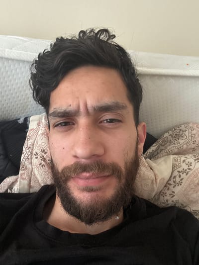 The image displays a close-up portrait of an adult male lying down, oriented horizontally on what appears to be a bed. He has dark, curly hair and a full, dark brown beard. He is wearing a black long-sleeved top, possibly a sweatshirt. His head rests on a pillow and is partially covered by a blanket, both adorned with a light brown and cream patterned fabric featuring a Paisley-like design.

The man looks directly at the camera with a neutral to slightly pensive expression, his eyebrows subtly furrowed. Visible skin details include a small red mark below his left eye and another on his forehead. The background shows a plain, light-colored wall on the left, and a white, textured fabric, possibly a curtain, towards the top right. The setting is indoors, likely a bedroom. No specific activity is depicted other than the individual resting. No text is visible in the image. The image provides no specific visual cues for time of day or weather, nor does it visually confirm the provided location context of Mid Devon, United Kingdom.
