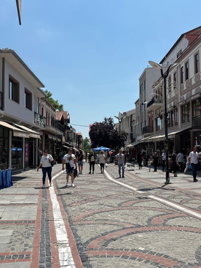 The image depicts a bustling pedestrian street in Edirne Merkez, Turkey, under a clear, bright blue sky. The street is lined with multi-story buildings featuring a blend of architectural styles, ranging from traditional designs with ornate balconies and pitched roofs to more modern structures with clean lines.

On the left side of the street, buildings are primarily two to three stories tall, displaying light-colored facades (white, cream) with brown window frames and dark awnings. Storefronts include "LAKE SHOES" and a partially visible "FUSS CAFE" sign. Some buildings further down have red-tiled roofs and traditional balconies.
On the right side, buildings also vary in height and style, exhibiting light grey, white, and brownish facades with multiple windows and balconies, often featuring decorative ironwork. Visible storefronts include "DİDE BOUTIQUE" and "FENOMEN."

The street itself is paved with a distinctive pattern. A central pathway runs down the middle, composed of grey rectangular stones flanked by red bricks, which in turn border a central white marble strip that shows signs of wear and cracking. On either side of this central strip, the pavement consists of grey and reddish-brown cobblestones arranged in concentric or curving patterns. Metal drainage grates and manhole covers are integrated into the paving.

Numerous pedestrians are present on the street, walking in both directions. Individuals are dressed in casual, light summer clothing such as t-shirts, jeans, and shorts, indicating warm weather. On the left, a woman in a white t-shirt and dark jeans walks with a man in a white t-shirt and denim shorts. Further along, groups and individuals are seen strolling, some carrying bags, suggesting shopping activity. A child walks alongside an adult on the right side of the street.

Street furniture includes tall, modern lampposts with distinctive disk-shaped lights, some featuring elaborate decorative ironwork. A dark-leaved tree with purple foliage provides shade over a small area on the left, where blue umbrellas are visible, likely covering outdoor seating. The bright sunlight casts distinct shadows from buildings and people onto the pavement.