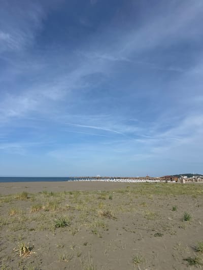 The image depicts a beach in Donji Štoj, Montenegro. The sky is blue with scattered clouds. The beach is sandy with sparse vegetation. White beach umbrellas are lined up along the shore. A building is visible in the background.