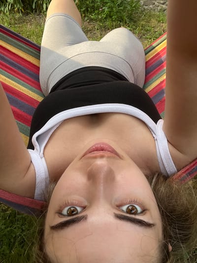A young woman is lying in a hammock outdoors, with grass and foliage visible in the background. She is wearing a black tank top with white trim and light gray athletic shorts. The hammock is made of striped fabric in red, yellow, green, and blue. The woman is looking up towards the camera, with a soft smile on her face, suggesting a relaxed and content mood. Her brown hair is spread around her head. The lighting suggests it is daytime, likely sunny, given the shadows and the overall brightness of the image. The presence of grass and greenery indicates a natural, outdoor setting. The image appears to be a selfie, taken from a high angle. There is no visible text in the image.