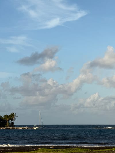 This image depicts a coastal scene in Māhina, France. The foreground shows a dark, possibly volcanic, sand beach with patches of green vegetation and small waves breaking. The mid-ground is dominated by a dark blue, choppy body of water with visible surface waves. A white sailboat, with its mainmast upright and sails furled, is moored or anchored in the water towards the left side of the frame. To the left of the sailboat, a small landmass extends into the water, featuring at least two prominent palm trees and a wooden pier or jetty. The background consists of a light blue sky with scattered white and grey cumulus and cirrus clouds. No individuals are present, and no active human interactions are depicted.