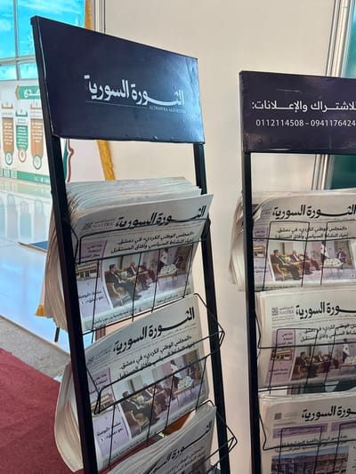 The scene is situated indoors, in Al-Ghizlaniya, Syria. Two black metal multi-tiered newspaper display racks are prominently positioned. The left rack features a top signboard displaying "الثورة السورية" (Al-Thawra Al-Suriyya) with the English transliteration "ALTHAWRA ALSUYRIYA." The right rack's signboard provides contact information for subscriptions and advertisements: "للاشتراك والإعلانات: 0112114508 - 0941176424." Both racks are fully stocked with multiple copies of "الثورة السورية" newspaper. The visible front pages uniformly feature a color photograph depicting several men in formal attire seated around a table during what appears to be a meeting, with a flag visible in the background. Arabic headlines on the newspaper include "المجلس الوطني الكردي" (The Kurdish National Council) "في دمشق" (in Damascus) and "إحياء النشاط السياسي وآفاق المستقبل" (reviving political activity and future prospects), along with text mentioning "NASTEX" and "ماذا تعني استعادة مطار القامشلي" (What does the restoration of Qamishli Airport mean?). The environment consists of a light-colored wall in the background. A red carpet is visible on the floor in the left foreground. To the left and rear, a pleated light-colored fabric and a green and white banner with circular graphics and Arabic text are partially discernible. No individuals are present in the frame.