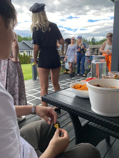 A group of people are gathered on a wooden deck outside a modern house. The sky is overcast with some patches of blue. In the foreground, a person is holding a tortilla chip, and a watch is visible on their wrist. A black wicker table is in front of them with bowls of food and a large water dispenser. In the middle ground, several women are dressed in costumes. One is wearing a black outfit and a beret, another a striped dress, and a third in blue overalls. In the background, a green lawn and more houses are visible. The scene suggests a casual outdoor gathering, possibly a party or barbecue, in a suburban residential area of Joensuu, Finland.