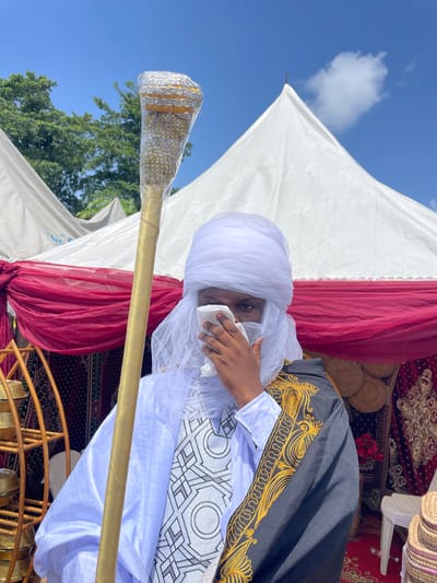 The media file shows a close-up of a person wearing traditional attire, likely at an outdoor event in Alagba, Nigeria. The individual is wearing a white turban and a white garment with intricate geometric patterns, over which a black robe with elaborate gold embroidery is draped. They are holding a white cloth to their face, partially obscuring their features, and their eyes are visible.
In the foreground, a decorative golden staff or scepter is held, adorned with silver and gold embellishments and what appears to be small, bead-like elements. The background features elements of a tent or canopy structure, with white and deep red fabric draping. Beyond that, glimpses of a patterned rug and woven baskets are visible, suggesting a celebratory or ceremonial setting. The clear blue sky with a single white cloud and the presence of lush green trees indicate it is daytime and the weather is fair. The overall impression is one of tradition, culture, and possibly a significant ceremony.