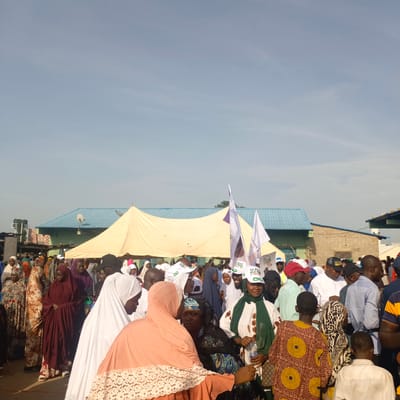 A medium outdoor shot depicts a daytime scene of a crowded gathering in what appears to be Gudi, Nigeria. A large beige tent sits in the background. The crowd is composed of people of various ages, predominantly of African descent, dressed in a mix of traditional and modern attire. Many women are wearing head coverings, including hijabs and traditional Gele headties. Several individuals are holding white flags with political messaging. There are buildings and structures visible behind the crowd and tent, including a light blue roofed building with a satellite dish. The weather appears fair with a partly cloudy blue sky.