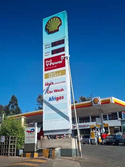 The image shows a Shell gas station in Gitaru, Kenya, located at latitude -1.226 and longitude 36.664. The gas station is open and operational. There are several vehicles parked, and several people are present, including some working at the gas station. The sign indicates that the station sells fuels unleaded, fuel-flow diesel, and Shell V-Power performance fuel. The sign also indicates that the station offers Shell Select and a car wash. The gas station is located in a rural setting with trees in the background. The sky is blue and cloudless. The signage features red and yellow details. The station has a concrete base and a large canopy covering the fuel pumps. An Afrigas sign is visible below the Shell Select sign. There is a green, rectangular sign with a white logo on it. There are also yellow and black striped markings on the pavement near the sign. There is a fence on the left side of the image.